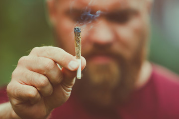 Sad or disappointed young man with beard is holding a smoking marijuana joint - legalization (or legalisation) medical cannabis concept - smoke is coming out of the weed or hashish cigarette