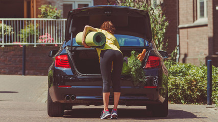 Young slim sporty woman in leggings taking a yoga mat, purchases (baguette bread and vegetables) and other things out of open car trunk (luggage van) - life and work balance, and good habits concept © Alina Rosanova