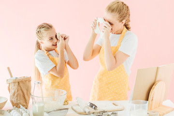 Obraz na płótnie Canvas happy mother and daughter looking at each other with dough pieces while preparing cookies isolated on pink