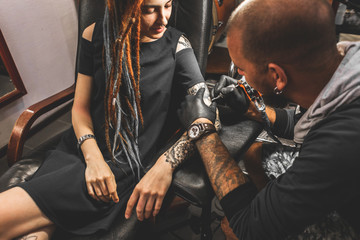 girl with dreadlocks in a tattoo parlor. The master creates a picture on the body of a young...