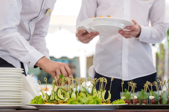 Two waiters serves banquet table in white marquee outdoor. Hands closeup