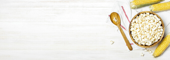 Brown wooden bowl with delicious traditional popcorn, fresh corn and a spoon on a light wooden background. Top view of a light meal background.