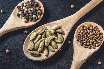 close-up view of wooden spoons with peppercorns, coriander seeds and cardamom on black