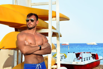hot muscular beach lifeguard in shorts with kayak and surf board standing in sand looking sexy with...