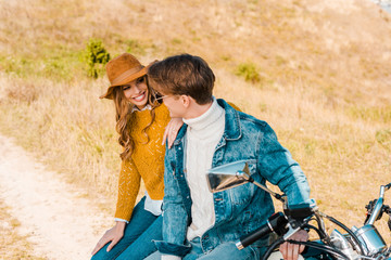 smiling couple sitting on motorbike and looking at each other on meadow