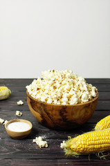 Brown wooden bowl with delicious traditional popcorn, fresh corn and a spoon on a dark wooden background. Top view of a light meal background.
