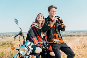 smiling boyfriend holding binoculars near girlfriend sitting on motorbike