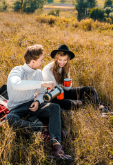 happy couple pouring hot drink from thermos and resting on rural meadow