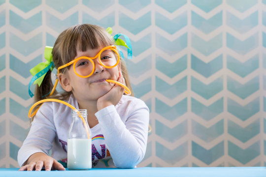 Little Smiling Girl Drinking Milk With Funny Glasses Straw On Blue Background. Growing Up Concept.