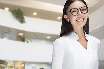 A young woman doctor smiling, standing in a hospital main hall. Communicate about doctors, hospitals, clinics, health care, medical and hospice care subjects