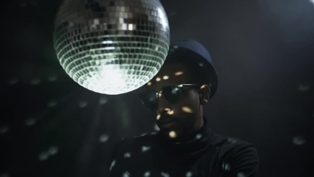 Low-key portrait shot of cocky black man in hat and sunglasses spinning disco ball and partying isolated on dark background