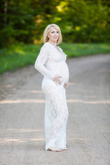 Beautiful pregnant woman in sheer long lacy white maternity dress looking dreamy on lonely road in forest