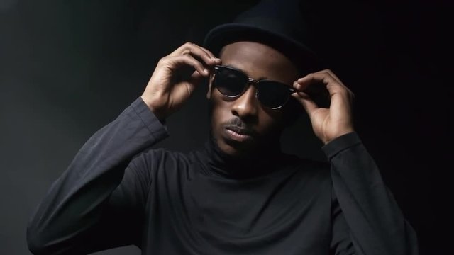 Low-key portrait shot of cocky black man in hat putting on sunglasses and posing isolated n dark background in foggy studio