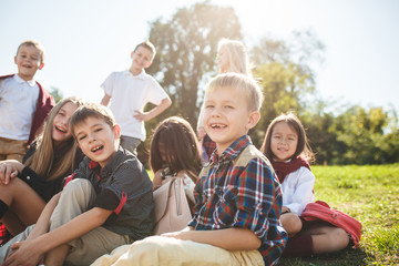 A group of happy smiling children of school and preschool age are sitting on the green grass in the park. The childhood, Kids fashion, school, education, friends, lifestyle, leisure, schoolchildren