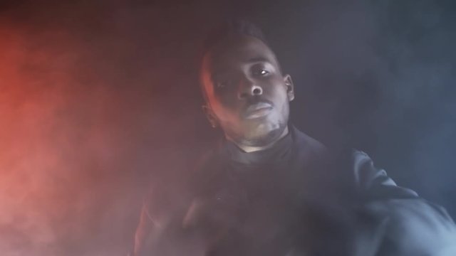Low-key portrait shot with red lighting: cool black man in jacket looking at camera and rapping in dark studio filled with smoke