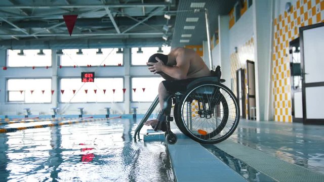 Disabled Man In A Wheelchair Putting On A Swimming Cap And A Goggles. Side Angle