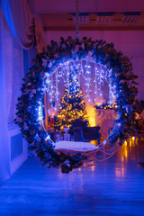 Large wreath of twigs and swing. Christmas evening by candlelight. classic apartments with a white fireplace, decorated tree, sofa, large windows and chandelier.