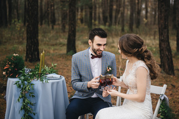 The bride and groom sit at a table set for two in the woods. They smile and drink mulled wine. Autumn. The concept of a romantic date
