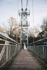 Fototapeta premium Happy young smiling bride and groom are standing on the suspension bridge. Wedding photos in an interesting place