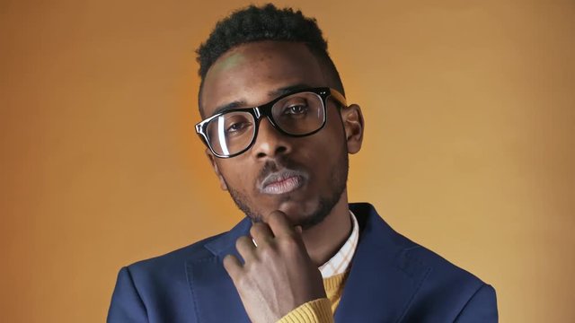Portrait shot of stylish arrogant black man in glasses and jacket holding hand to his chin and looking at camera while posing in studio with orange background