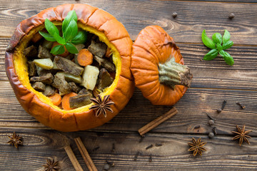 Baked meal in pumpkin on rustic brown wooden background. Stewed potatoes with carrot and roasted meat pork, decorated with basil leaves. Autumn mood, thanksgiving fall background.
