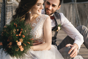 Happy young smiling bride and groom are sitting on the suspension bridge. Sunny wedding photos in...