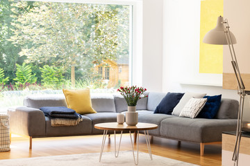 Flowers on table next to grey corner sofa with cushions in apartment interior with window. Real photo