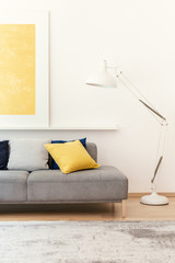 White lamp next to grey couch in modern living room interior with yellow poster and carpet. Real photo