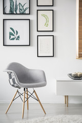 Grey armchair next to cupboard in white living room interior with gallery of posters. Real photo