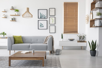 Wooden table in front of grey settee in natural living room interior with blinds and gallery. Real...