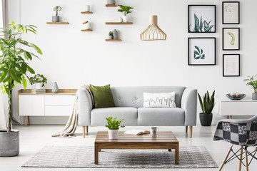 Flowers on wooden table in front of grey sofa in scandi flat interior with posters and armchair. Real photo - obrazy, fototapety, plakaty