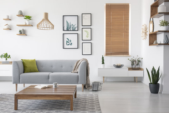 Wooden table in front of grey settee in natural living room interior with blinds and gallery. Real photo