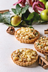 Homemade sweet apple shortbread tartlets with cinnamon sticks, walnuts, apples red leaves branches above on white marble table. Autumn baking.