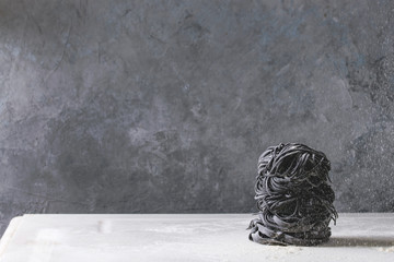 Variety of italian homemade raw uncooked cuttlefish ink black pasta spaghetti and tagliatelle in stack with sprinkling semolina flour on white marble table. Copy space.