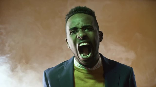 Portrait shot with green lighting: angry black man standing isolated on orange background in studio filled with smoke and screaming before camera