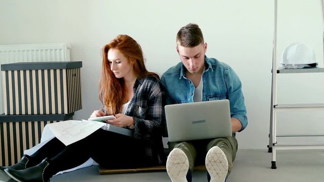 Young couple talking and using tablet, laptop and blueprints on floor at their new home
