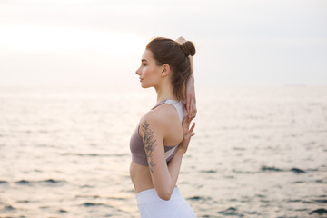 Fototapeta na wymiar Beautiful girl in sporty top and white leggings holding hands behind back with sea view on background. Young woman dreamily looking aside while practicing yoga by the sea