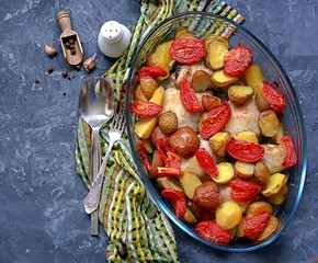 Baked chicken with potatoes and tomatoes in a glass form for baking on a dark gray background. Top...