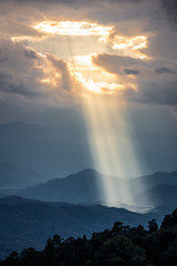 Obraz na płótnie Canvas Bright sunlight shining through hole of clouds to dark scene of mountain range before sunset in Thailand rainforest area.