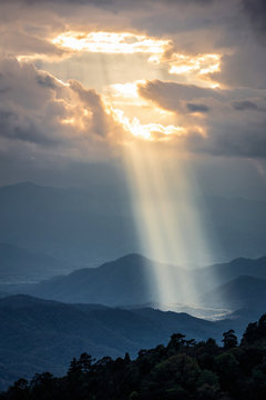 Fototapeta Bright sunlight shining through hole of clouds to dark scene of mountain range before sunset in Thailand rainforest area.