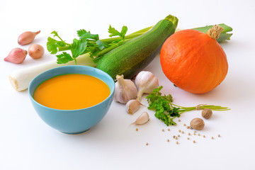 Turquoise bowl with pumpkin soup and raw ingredients, spice and herbs. Hokkaido pumpkin, onion shallots, leeks, celery, zucchini, garlic, parsley, nutmeg and white pepper, white plain background.