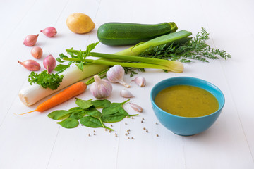 Turquoise bowl with spinach soup and raw ingredients, spice and herbs. The spinach leafs, onion shallots, leeks, celery, zucchini, garlic, parsley, carrot and white pepper, white wooden background.