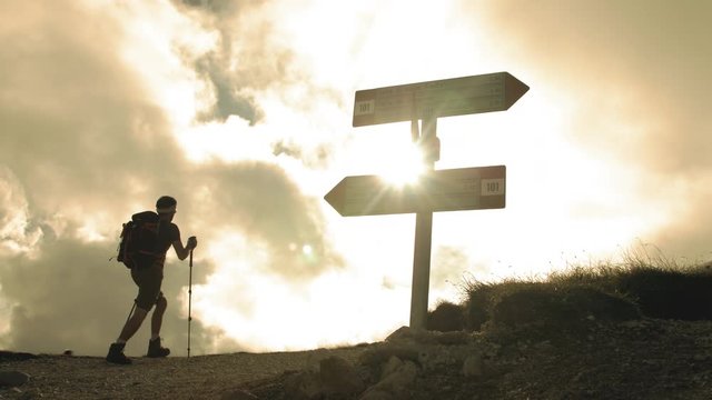 Trekker at mountain crossroad with signs walking up with backpack, boots and trekking poles. Clouds moving on the background. Abruzzo Italy.