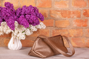 a bouquet of lilac and a female bag lie on a wooden table on a brick background