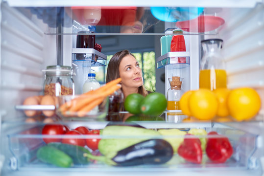 Confused hungry woman holding hands on head while standing in front of opened fridge full of groceries. Picture taken from the iside of fridge.