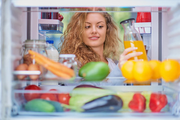 Woman standing in front of fridge full og groceries and taking juice. Picture taken from inside of...