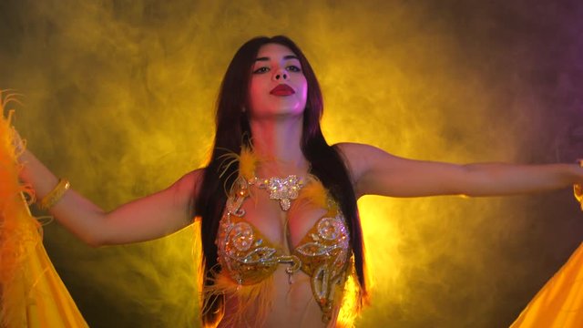 Seductive sexy traditional oriental belly dancer girl dancing on yellow neon smoke background. Woman in exotic costume with feathers sexually moves her semi-nude body.