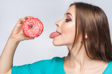 Side view profile of cheerful young beautiful girl in blue blouse standing, holding and showing pink donut, trying to suck with tongue out in grey background. Indoor, isolated, copy space, studio shot © khosrork