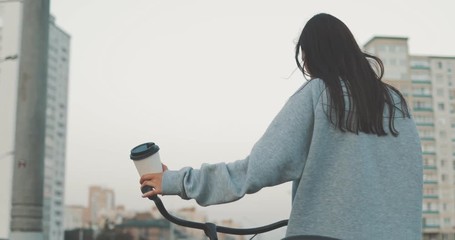 Young woman walking on city streets with bicycle and drinking coffee to go, sunshine, 4K video shooting by handheld gimbal