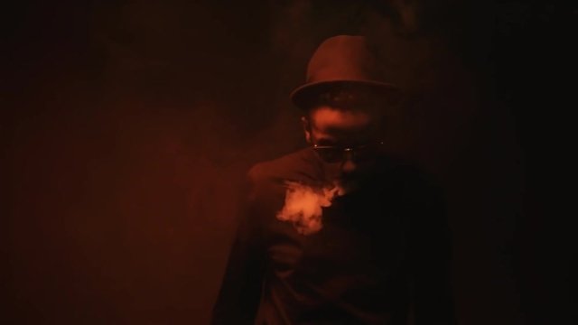 Low-key portrait shot with red lighting: confident black man in sunglasses and hat standing in dark studio and vaping, then exhaling clouds of smoke and making smoke rings